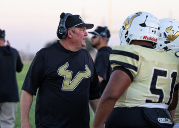 SHS-VHS-FB-29 | Press Pass Sports Football coach in a black shirt and headset talks to players in gold uniforms on the sideline during practice.