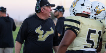 Football coach in a black shirt and headset talks to players in gold uniforms on the sideline during practice.