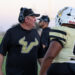 SHS-VHS-FB-29 | Press Pass Sports Football coach in a black shirt and headset talks to players in gold uniforms on the sideline during practice.