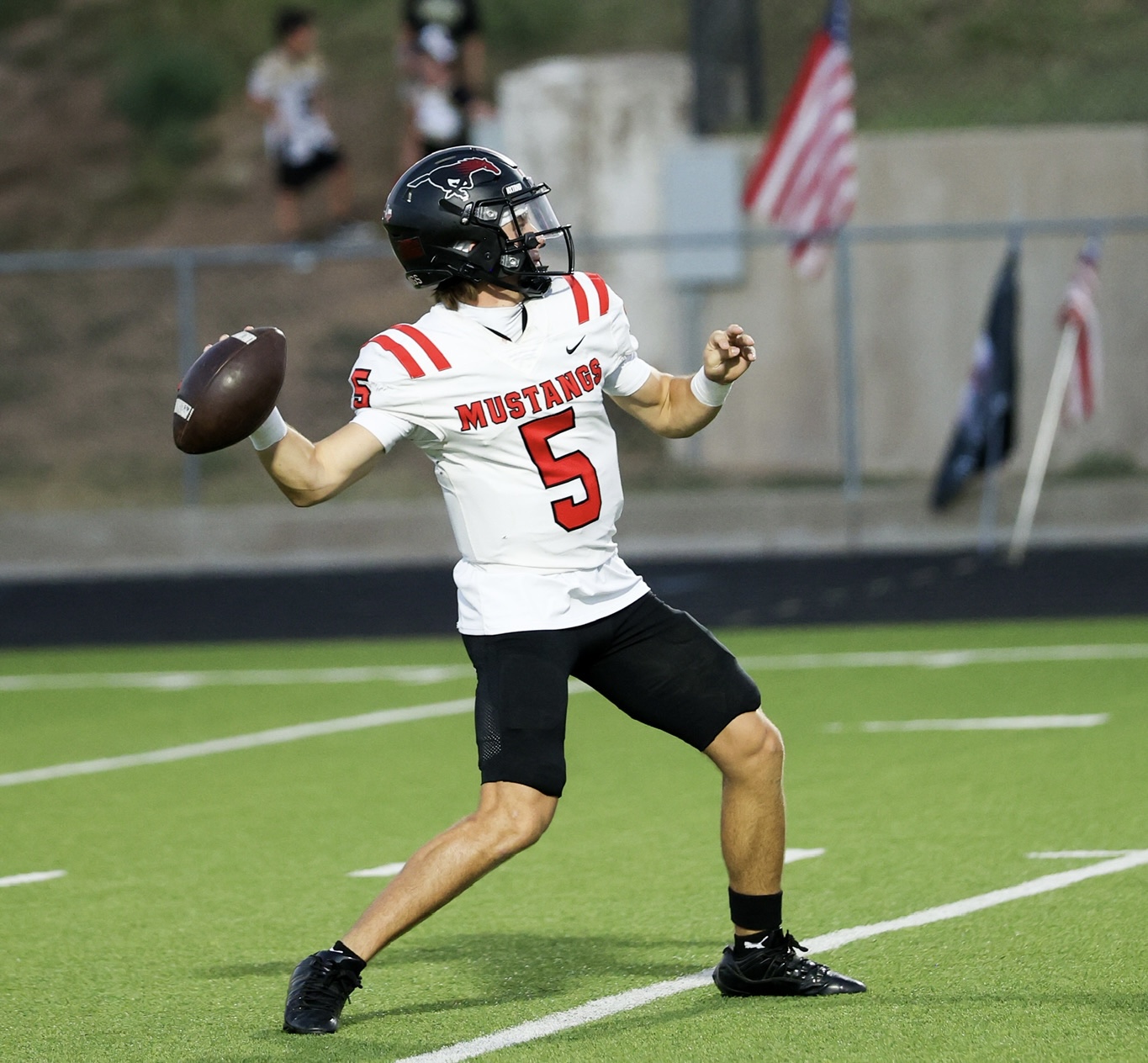 Football quarterback in a white Mustang jersey number 5, mid-throw on a green field with a football in his left hand.