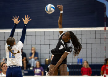 RHS-WPHS-VB-7 | Press Pass Sports Player in a black jersey jumps to spike the ball over the net as a defender in white reaches up to block the shot.
