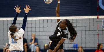 Player in a black jersey jumps to spike the ball over the net as a defender in white reaches up to block the shot.