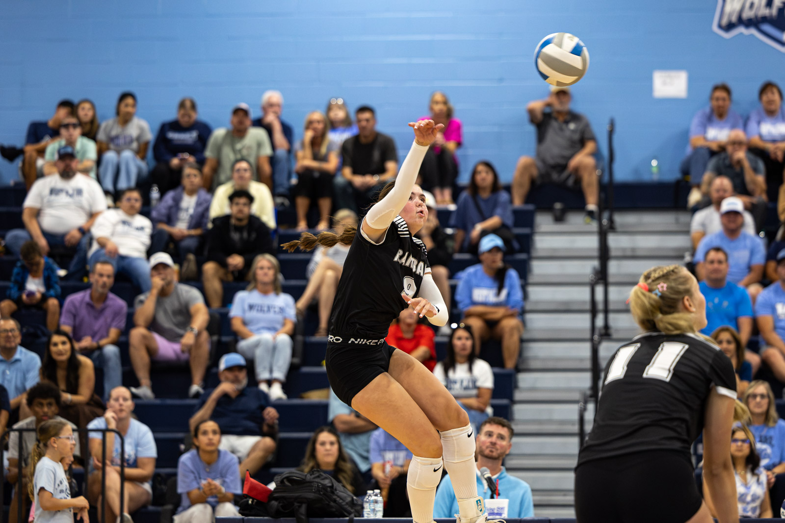 RHS-WPHS-VB-21 | Press Pass Sports Volleyball player in mid-air, reaching for the ball as spectators watch from blue bleachers behind her.