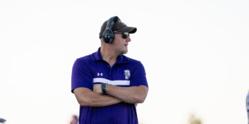 Middle-aged man wearing a purple polo shirt, cap, and sunglasses with over-ear headphones, arms crossed outdoors.