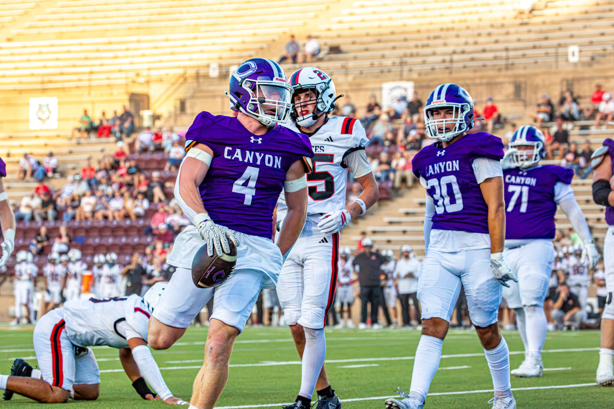 _v4a2811 | Press Pass Sports Canyon football player in purple jersey runs with the ball as teammates and opponents pursue on a sunny stadium field.