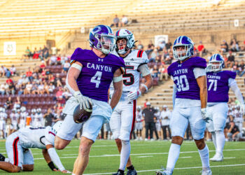 _v4a2811 | Press Pass Sports Canyon football player in purple jersey runs with the ball as teammates and opponents pursue on a sunny stadium field.