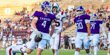 Canyon football player in purple jersey runs with the ball as teammates and opponents pursue on a sunny stadium field.