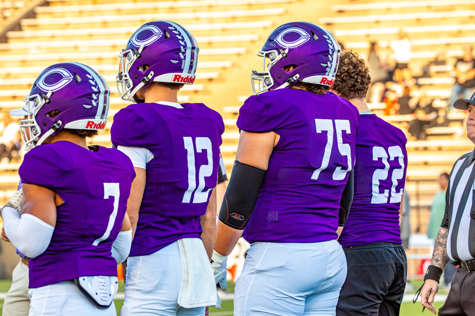 _V4A2693 | Press Pass Sports Group of football players in purple uniforms standing with backs to camera on the field, numbers 7, 12, 75, and 23 visible with helmets on.