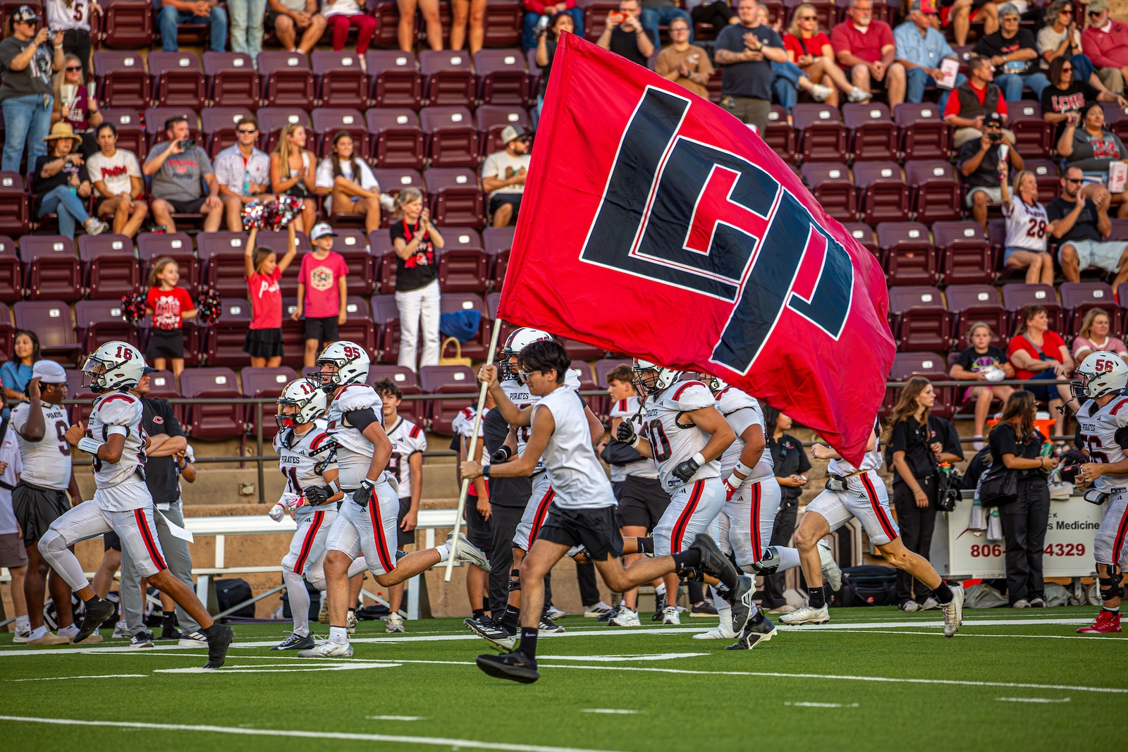 _V4A2705 | Press Pass Sports Football players sprint onto the field as a large red flag with a bold logo waves in the foreground, with fans in the stands behind them.