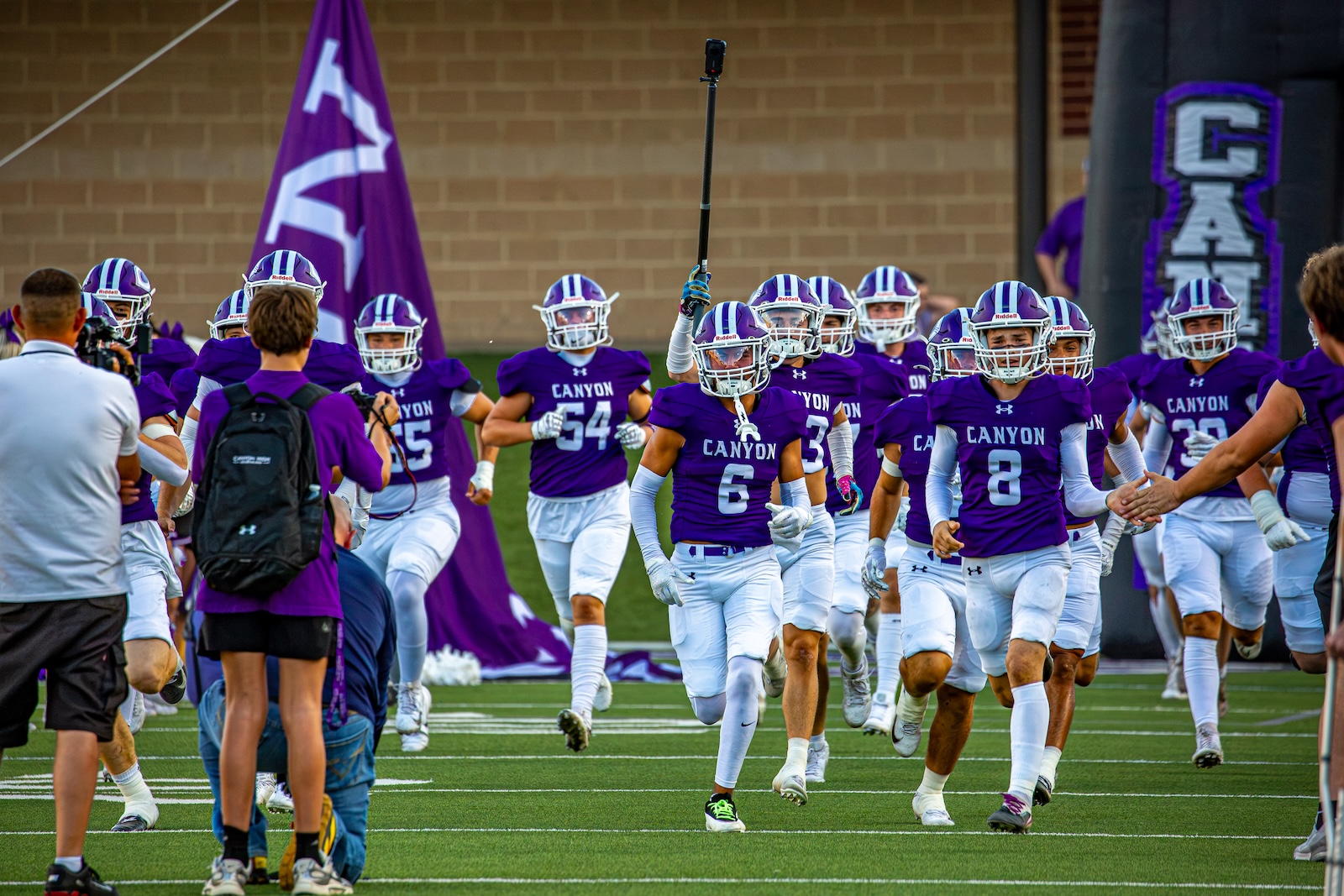 _V4A2709 | Press Pass Sports Canyon football players in purple uniforms run onto the field together, raising a coach or flag in the air as photographers capture the moment.