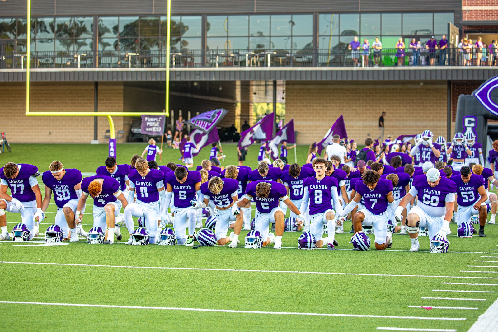 _V4A2720 | Press Pass Sports Canyon football team kneeling on a green field in purple jerseys and white pants, with helmets on the turf and fans in purple in the background.