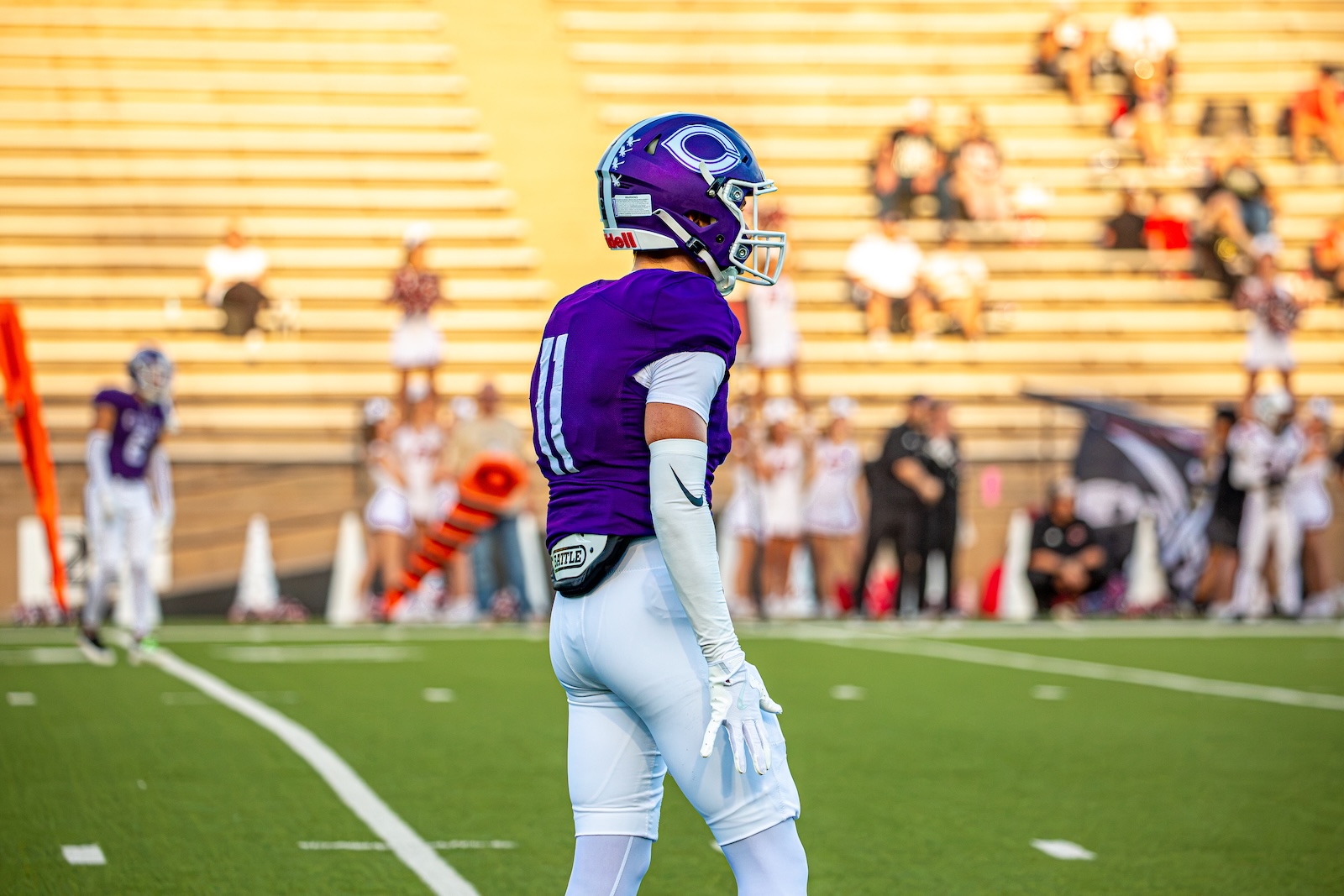 _V4A2727 | Press Pass Sports American football player in a purple jersey and white pants stands on the field, helmet on, during a game with bleachers in the background