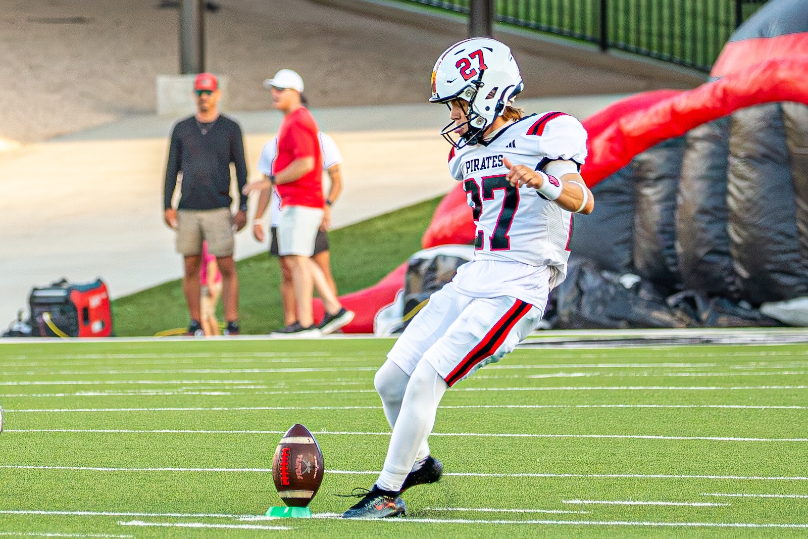 _V4A2728 | Press Pass Sports Football kicker in white Pirates uniform (number 27) about to kick a ball from a tee on a green field, with coaches in the background
