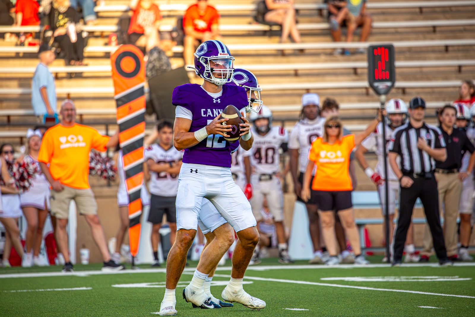 _V4A2738 | Press Pass Sports Football player in purple jersey (number visible) holds the ball at chest level on a green field, preparing to throw, with teammates and spectators behind him.