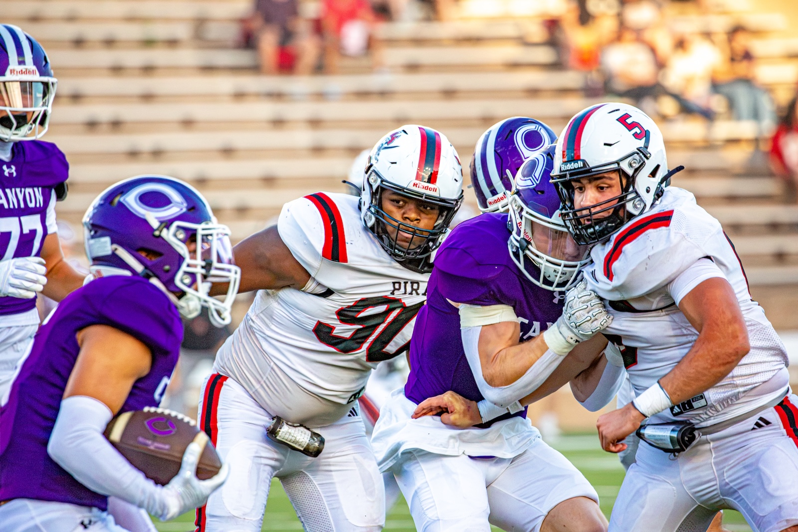 _V4A2746 | Press Pass Sports Football game action: players in purple and white uniforms crowding the ball carrier as a tackler grips his opponent close, on a sunlit field with empty bleachers in the background.