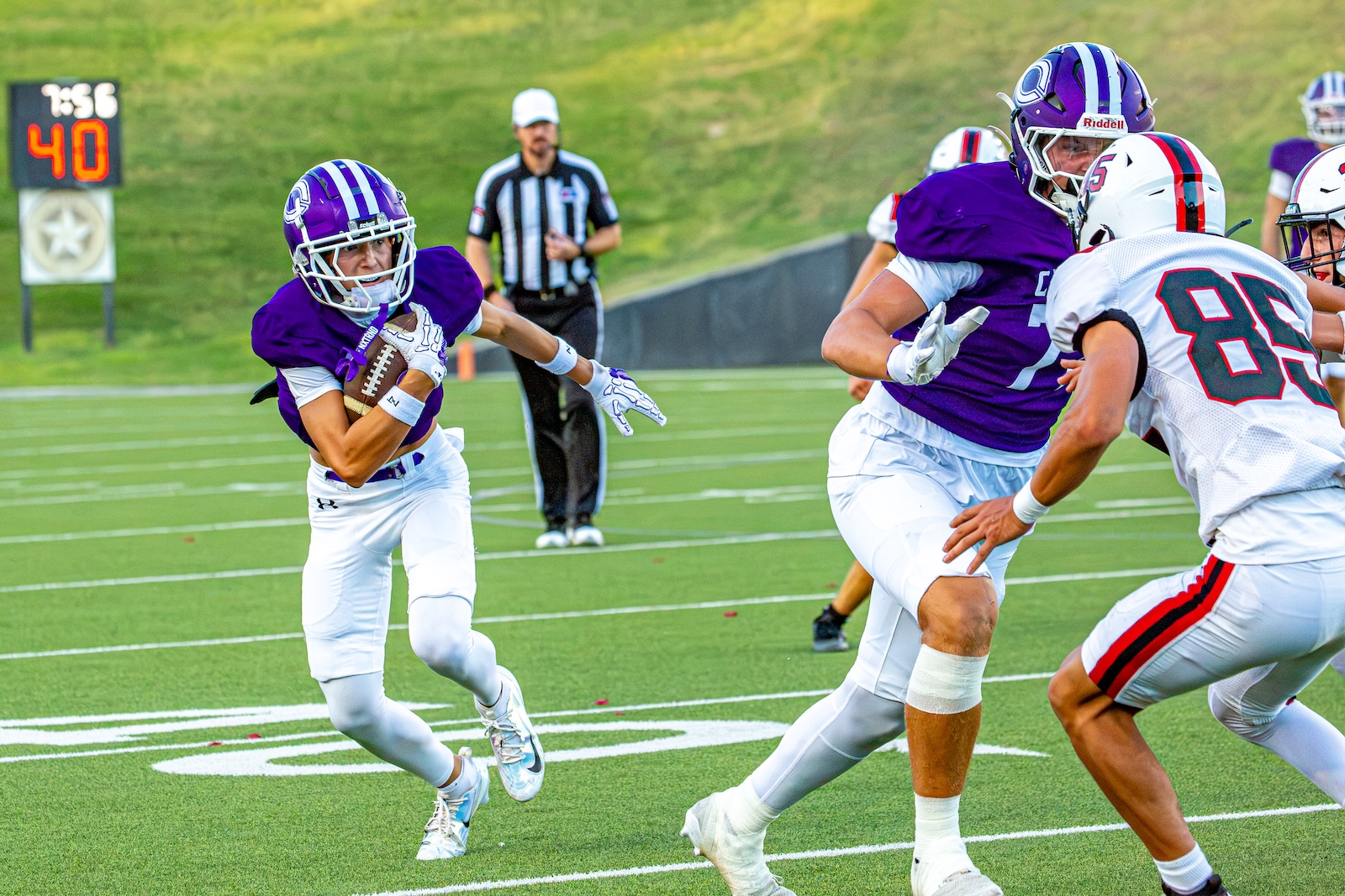_V4A2778 | Press Pass Sports Football players in purple jerseys chase the ball on a green field; a running back_takes a handoff while others block nearby.