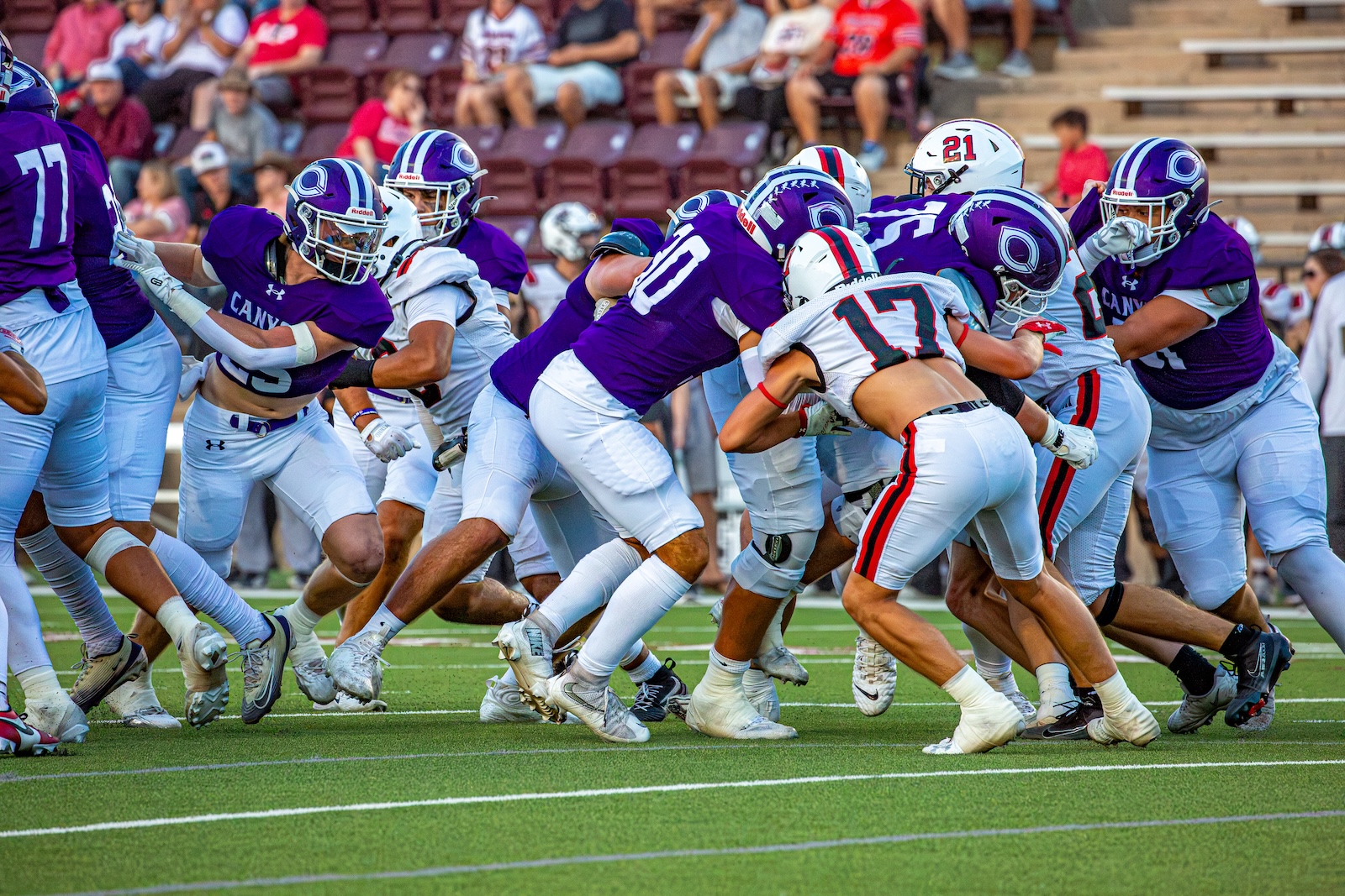 _V4A2793 | Press Pass Sports American football players in purple and white jerseys clash at the line of scrimmage on a green field, with spectators in the stands behind them.