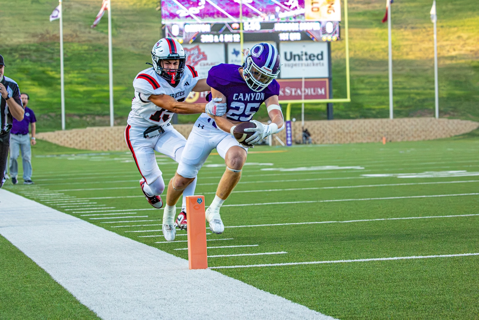_V4A2807 | Press Pass Sports Purple-jersey football player (Canyon) reaches for the ball near the orange pylon as a defender in white tries to tackle him on the sideline field edge.