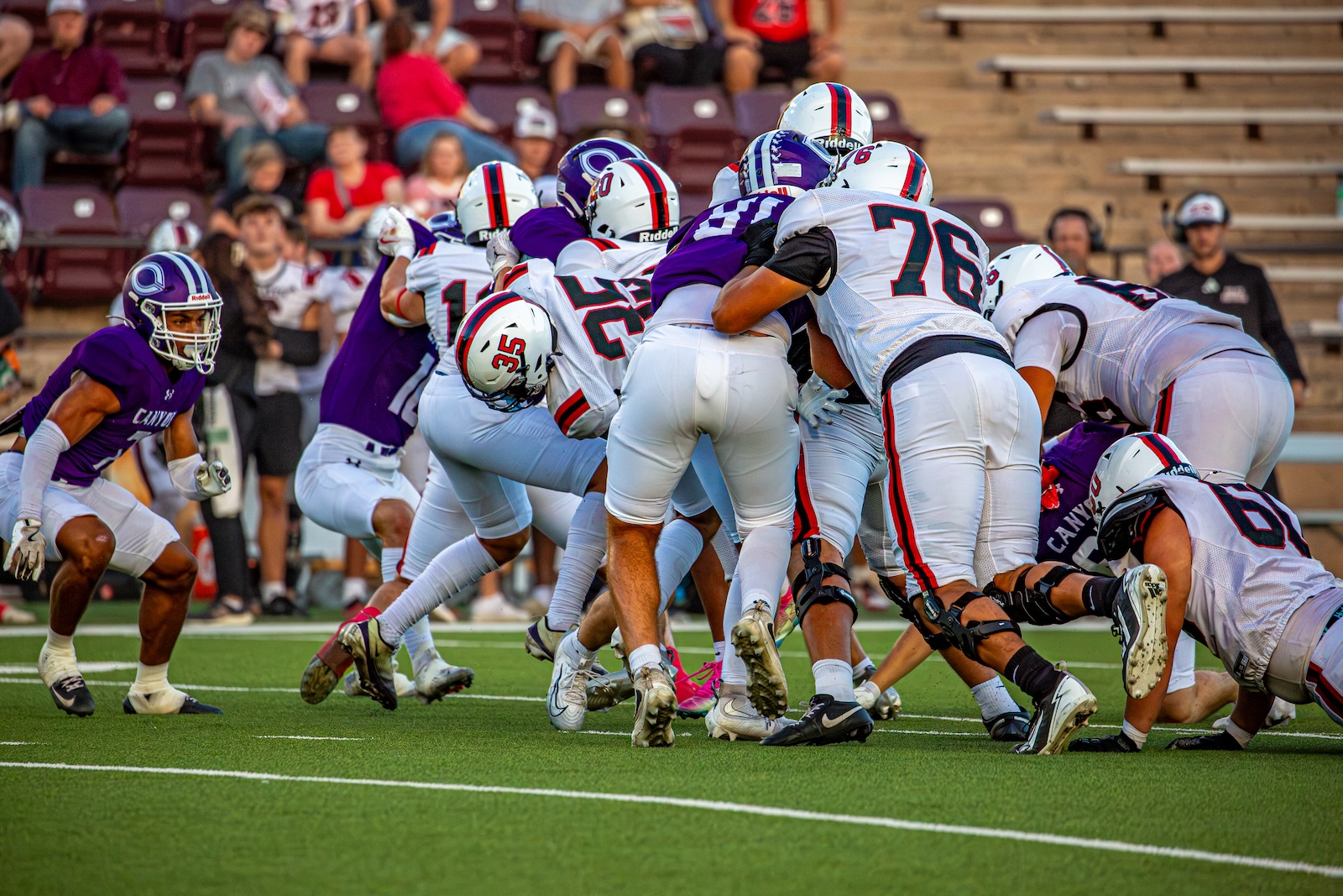 _V4A2847 | Press Pass Sports Group of football players in white and purple collide at the line of scrimmage on a green field with stands in the background, mid-play.