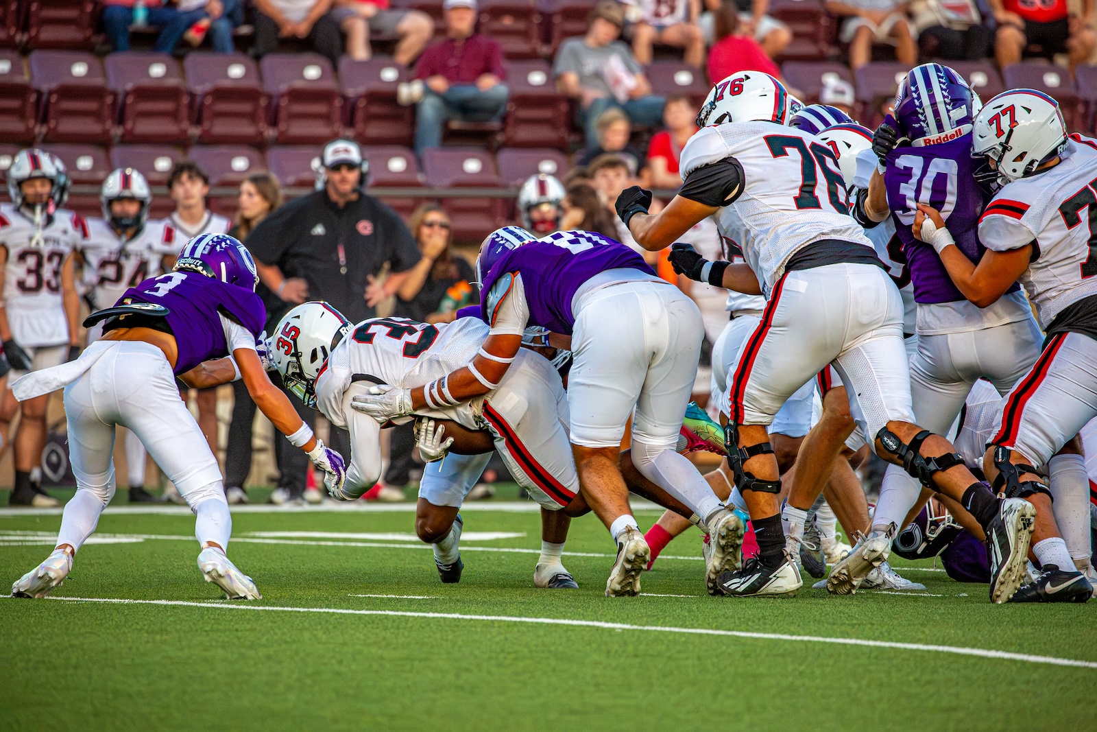 _V4A2848 | Press Pass Sports Football players in white and purple uniforms collide at the line of scrimmage on a green field with a crowd in the stands behind them earlier in the playtime.