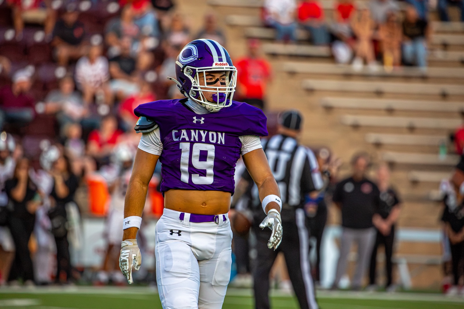_V4A2851 | Press Pass Sports Canyon football player #19 in a purple jersey and white pants on the field during a game, crowd in the stands behind him.