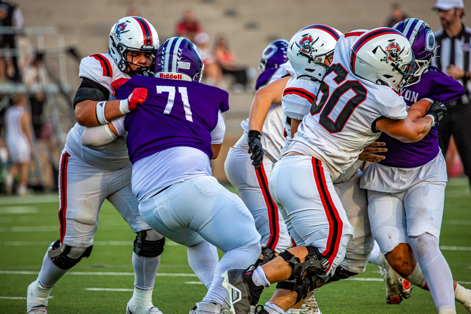 _V4A2879 | Press Pass Sports Football players in white and purple uniforms tackle each other on a green turf field during a game, with several players pushing and grabbing at one another.
