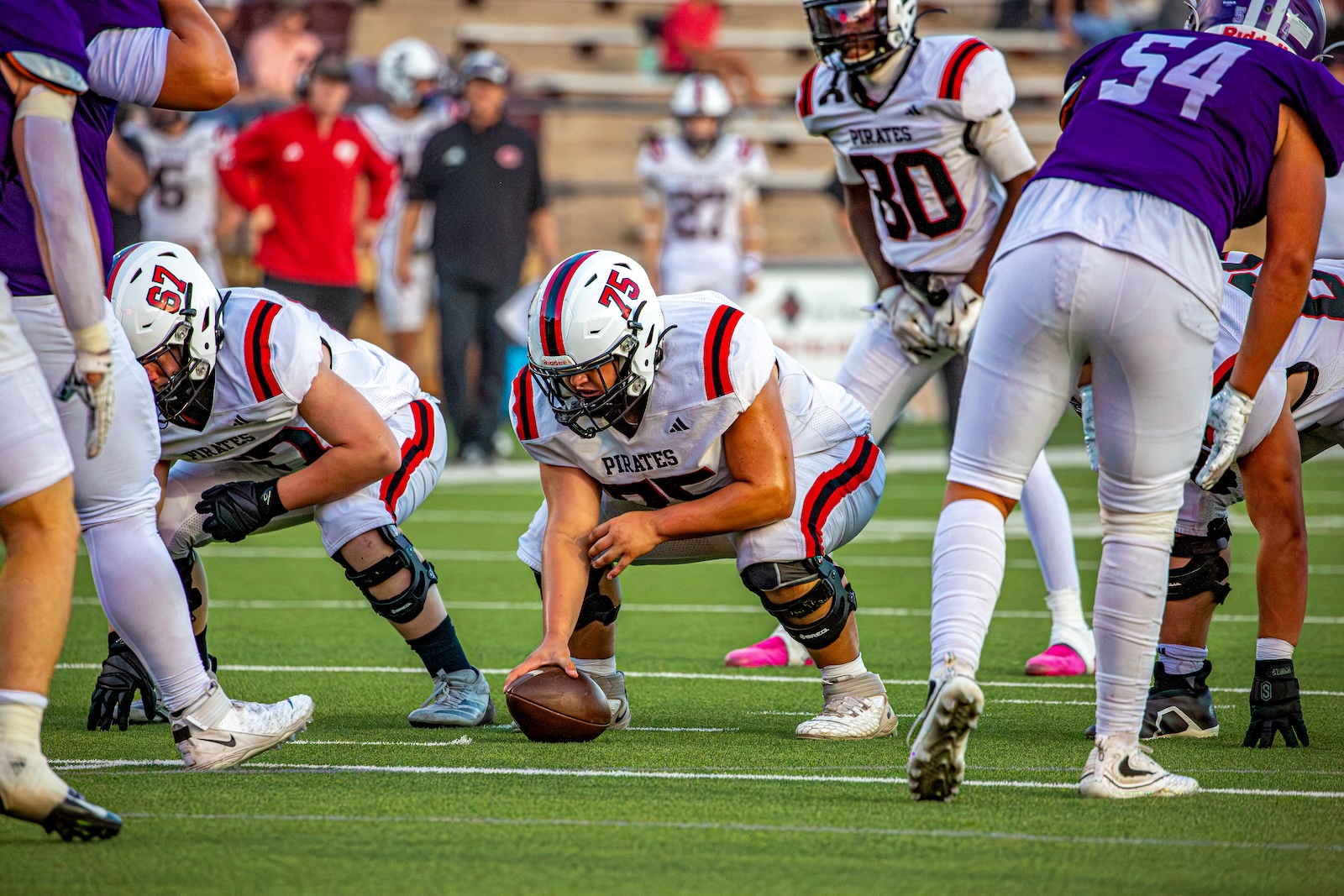 _V4A2889 | Press Pass Sports Pirates football center snaps the ball from a low stance amid linemen on a green turf field
