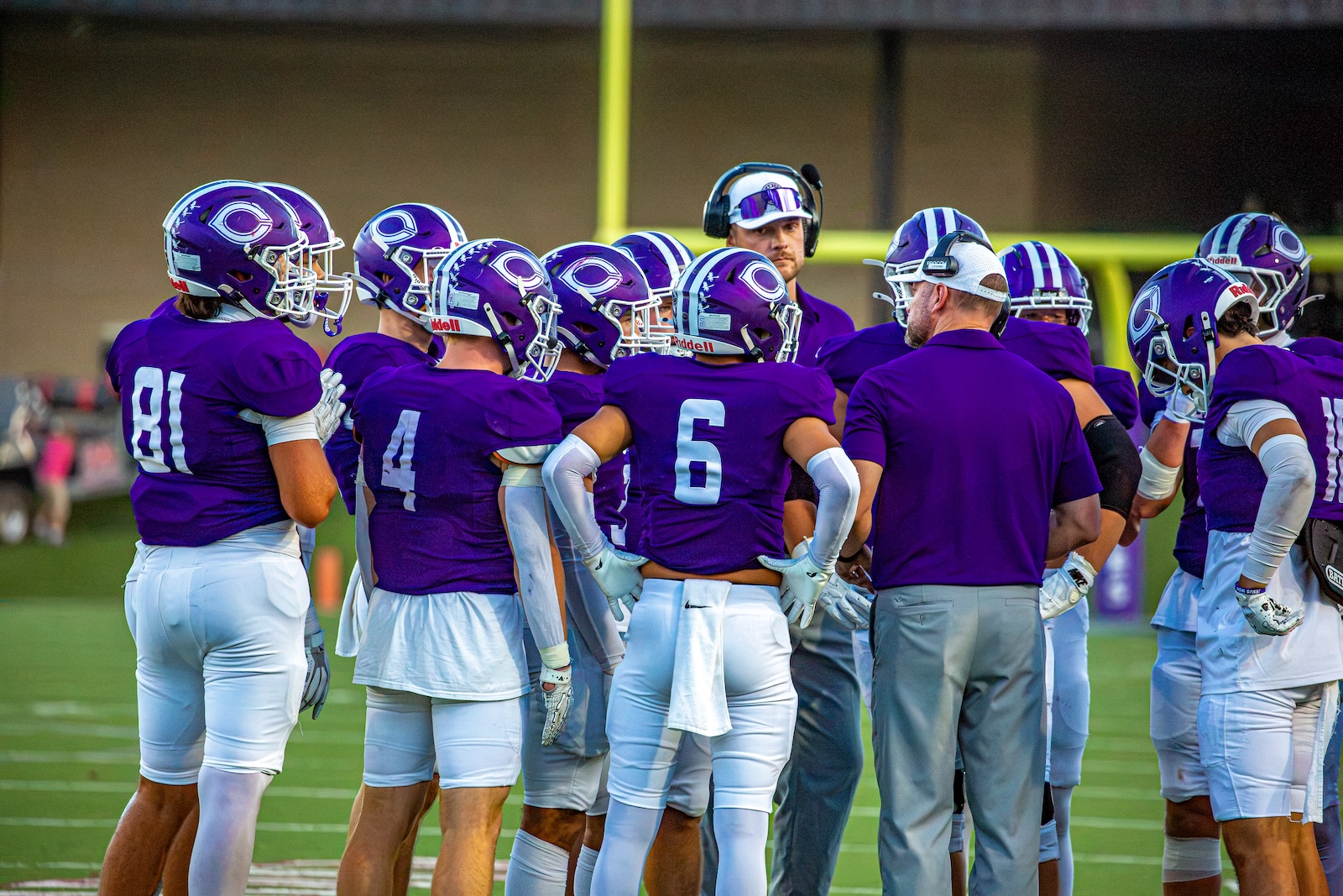 _V4A2917 | Press Pass Sports Purple-clad football players huddle with coaches on a green field before a game.
