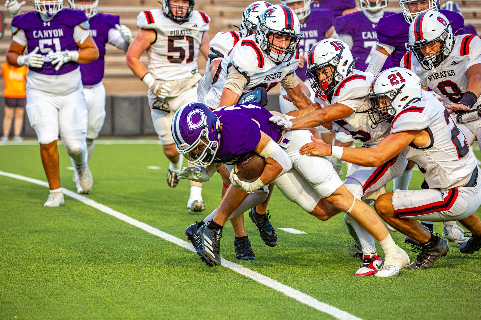 _V4A2973 | Press Pass Sports Purple-clad running back is tackled by Pirates players in white uniforms on a green football field.