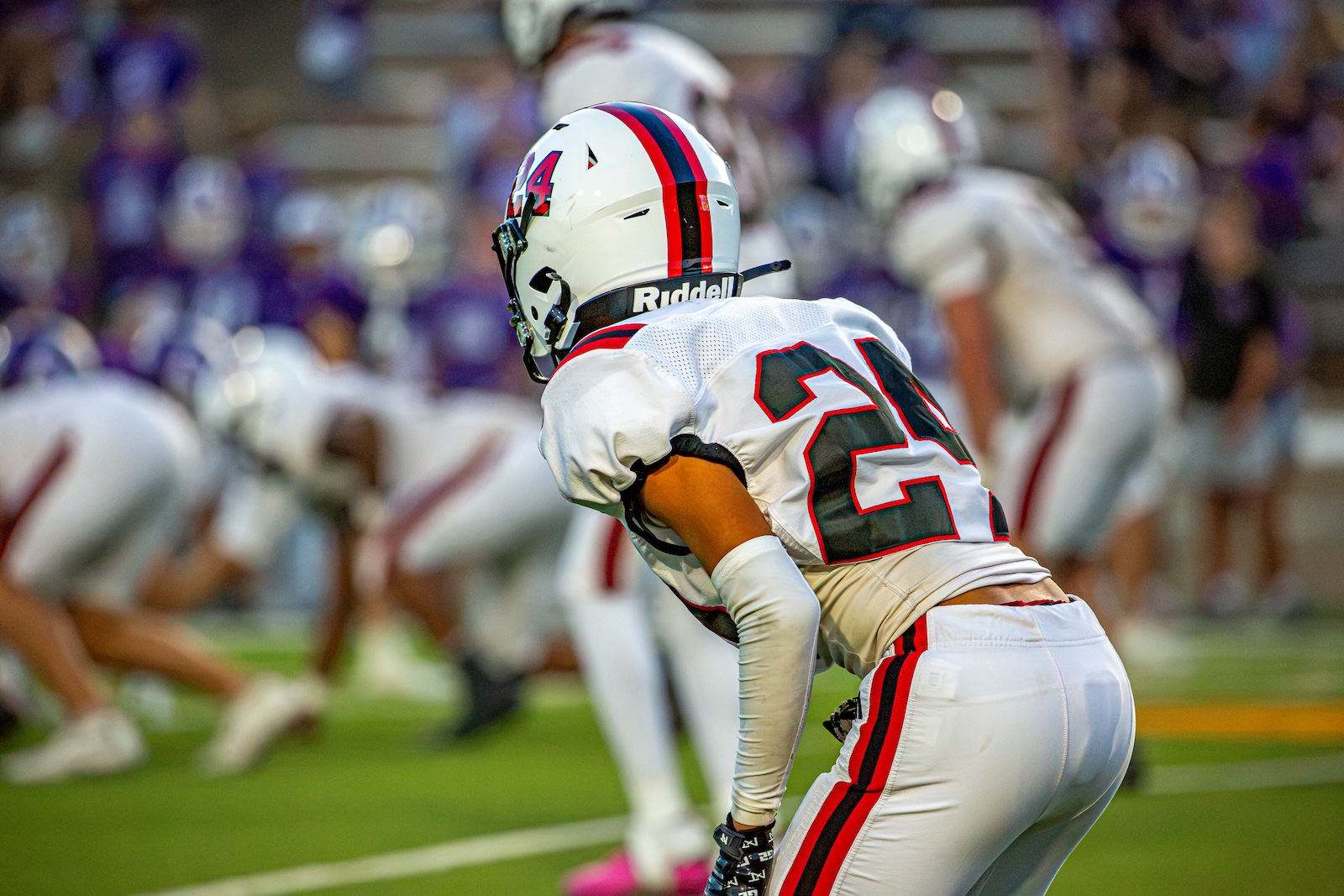 _V4A3019 | Press Pass Sports Football player in white jersey #24 crouches in a defensive stance on a green football field, helmet with red and blue stripes visible.