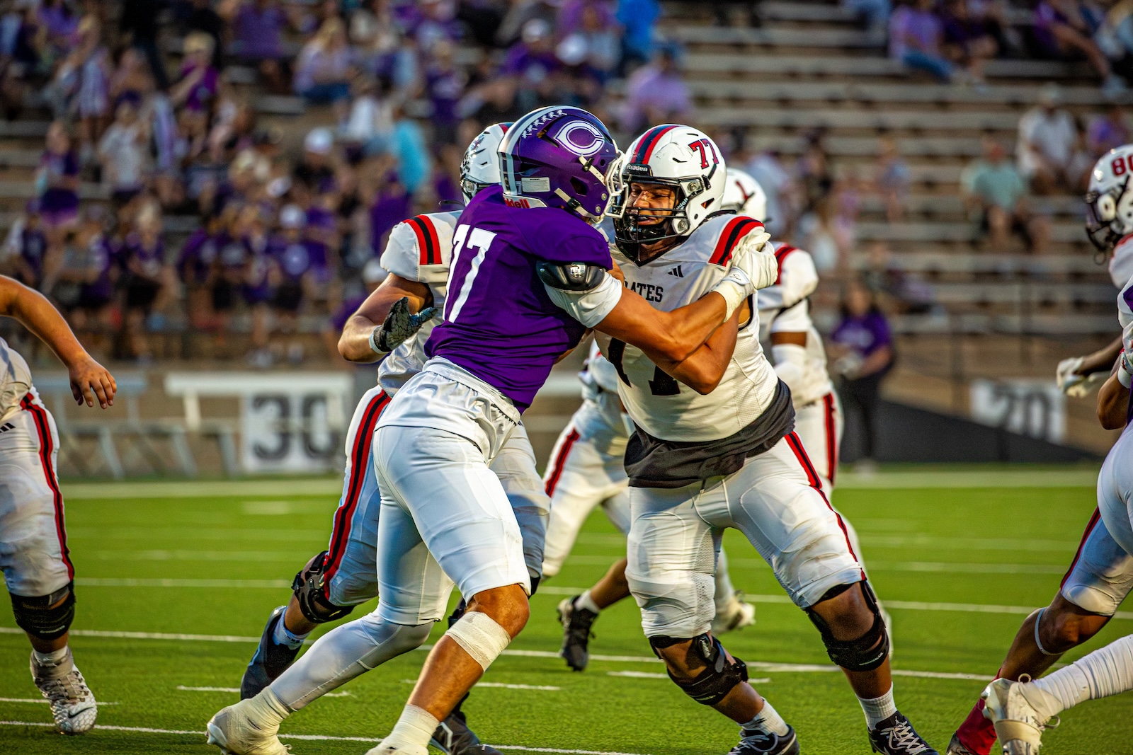 _V4A3058 | Press Pass Sports Two football players collide as a purple-jerseyed defender (#17) blocks a white-jersey opponent, on a green field with a crowded stands backdrop.