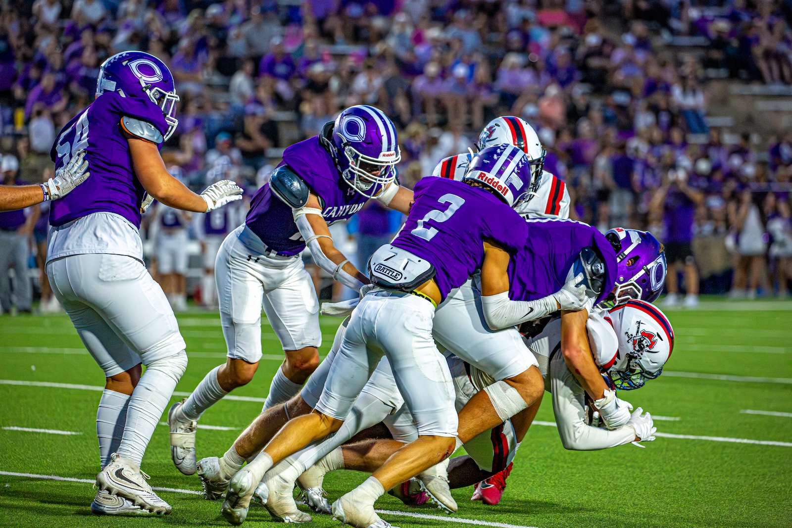 _V4A3089 | Press Pass Sports Group of football players in purple jerseys tackle near the sideline on a bright green field as fans watch from stands.