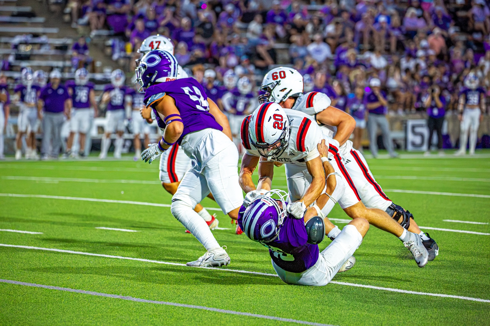 _V4A3094 | Press Pass Sports Football game action: a purple-clad defender pursues a ball carrier as a tackle unfolds on a bright green field with fans in the stands behind.
