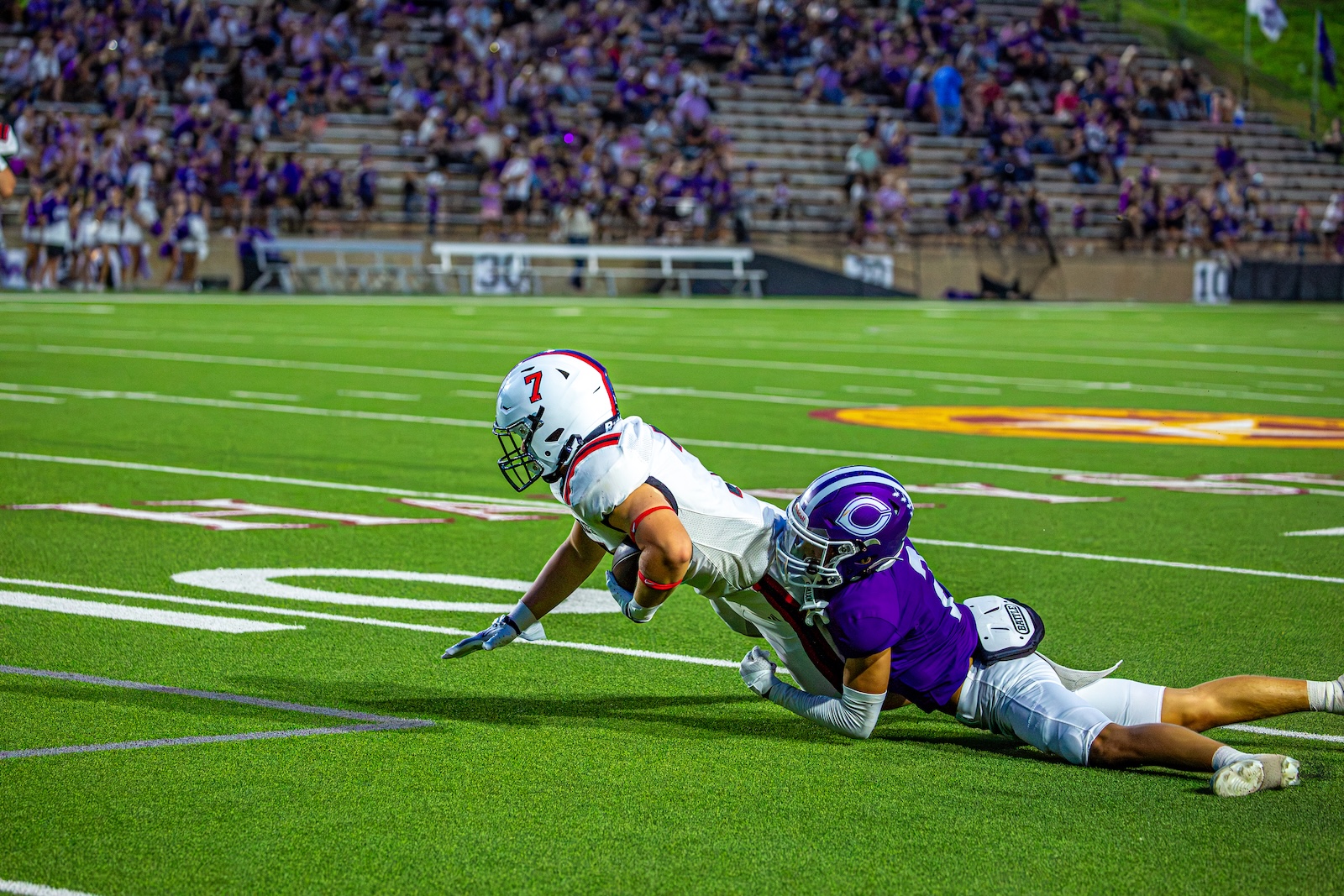 _V4A3109 | Press Pass Sports Football player in white dives forward while a purple-clad defender grabs at his legs on a bright green field with a packed stands backdrop.