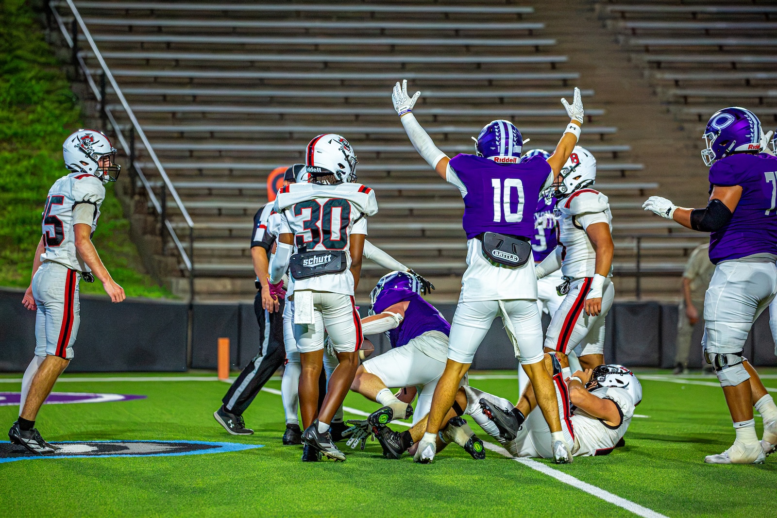 _V4A3171 | Press Pass Sports Football players in white and purple uniforms surge around a downed ball on a green turf, one team celebrating with raised arms as others regroup nearby in a stadium.