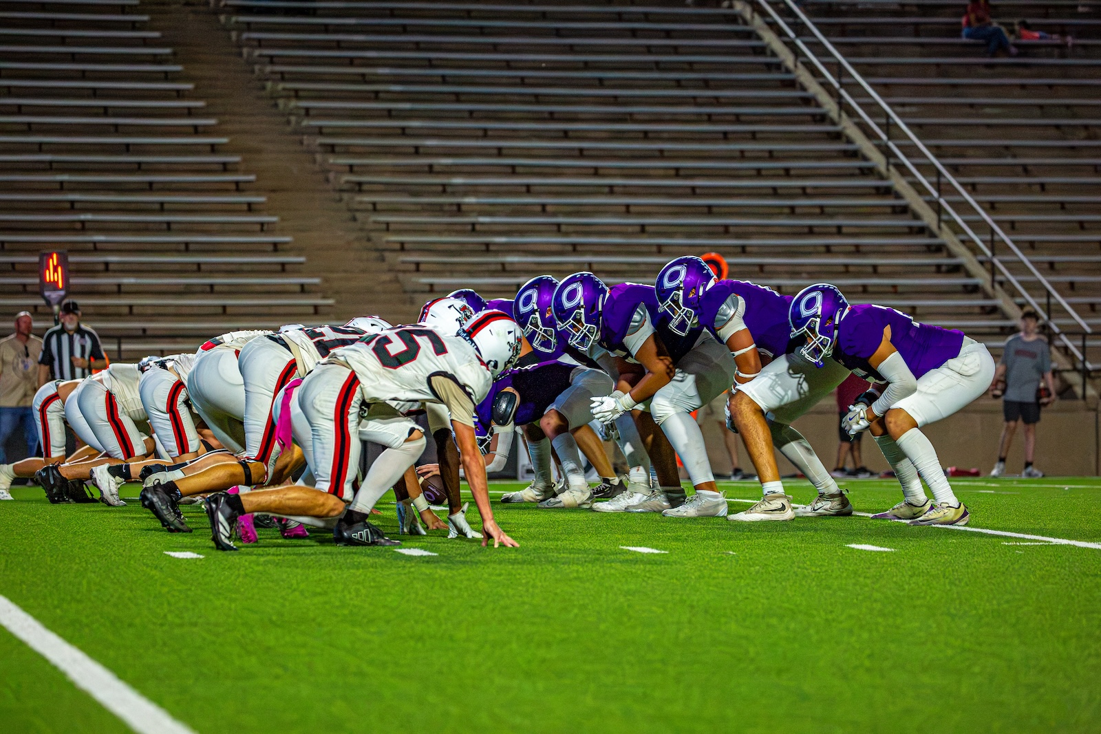 _V4A3177 | Press Pass Sports Two football teams line up at the line of scrimmage on a bright green field, white helmets facing purple helmets before snap.