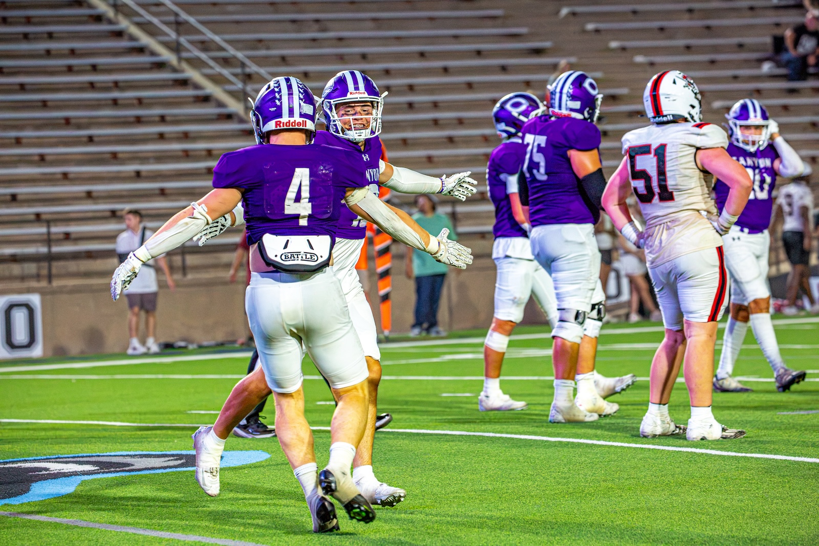 _V4A3320 | Press Pass Sports Football players in purple jerseys celebrate on a green turf field after a play, their arms outstretched in a cheer.