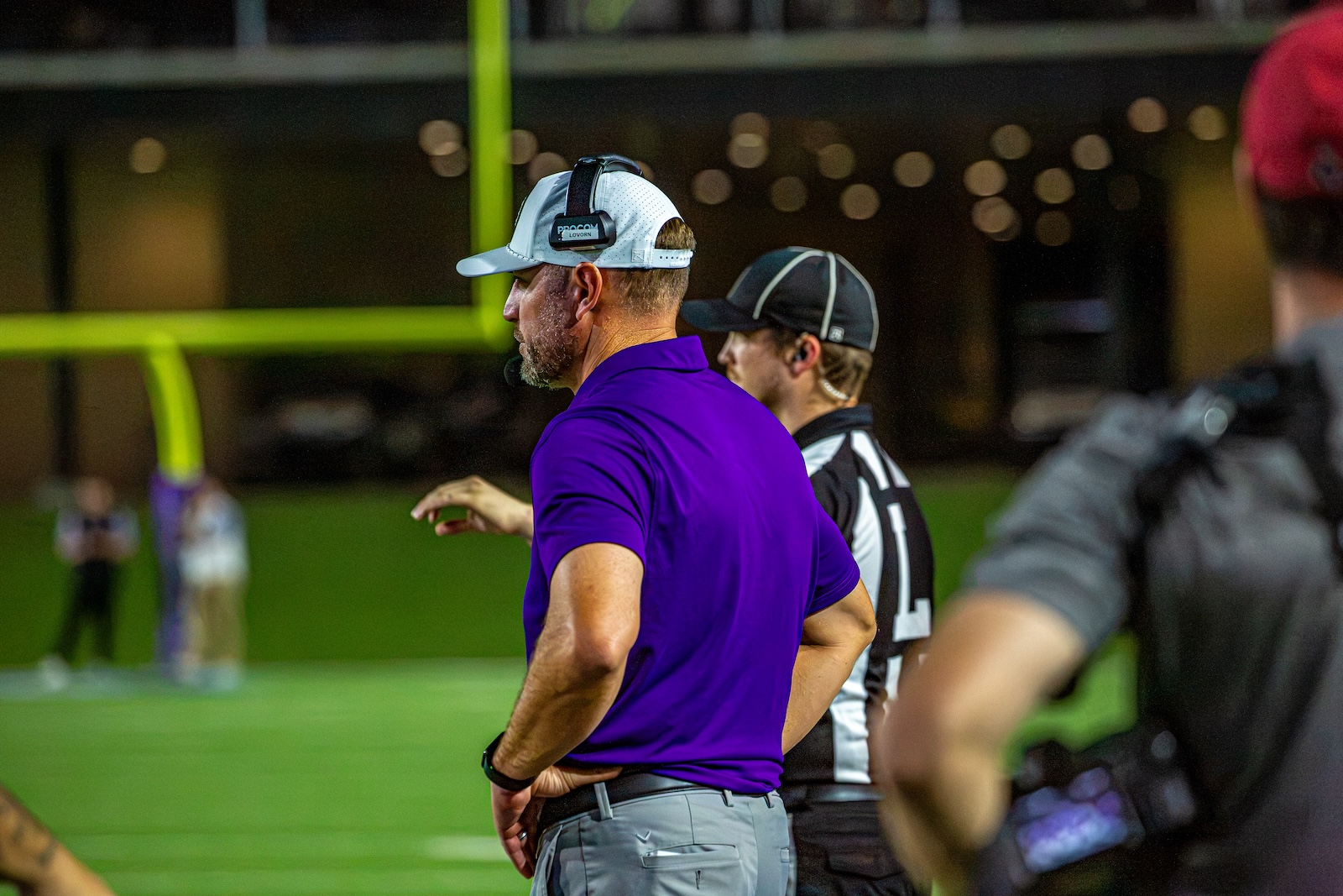 _V4A3331 | Press Pass Sports Coach in a purple polo and white cap with headset stands on the sideline of a football field.
