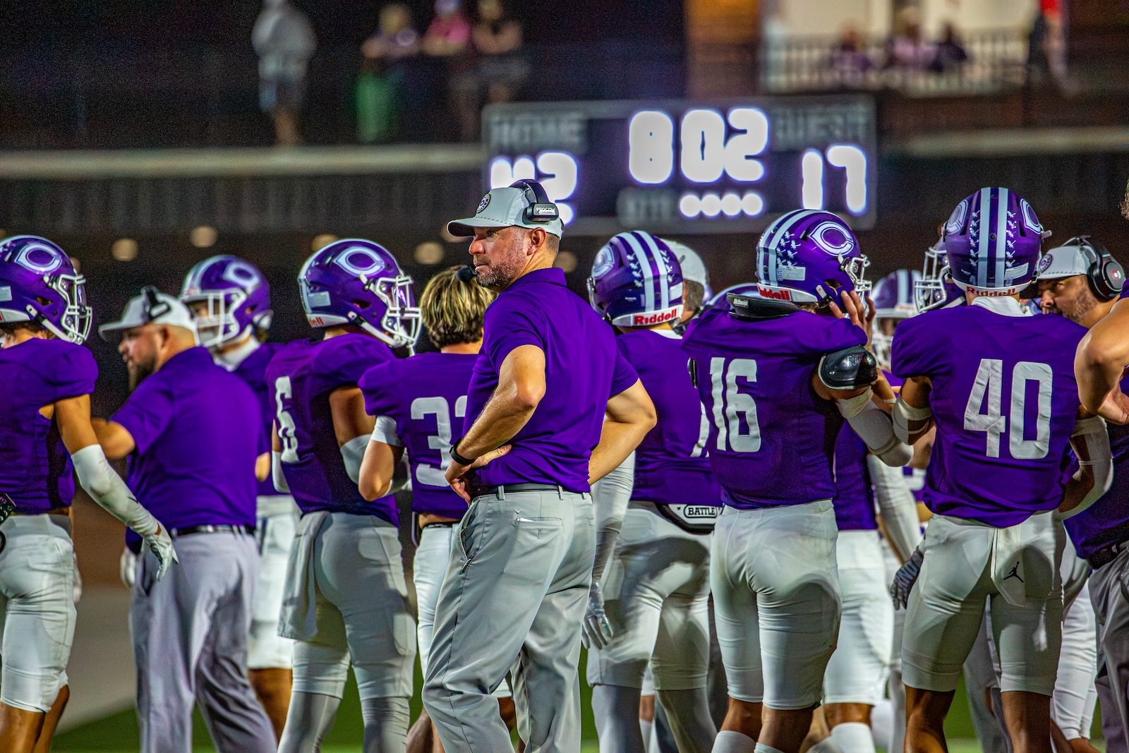 _V4A3414 | Press Pass Sports Football coach in a purple shirt and cap stands with his purple-clad team on the sideline during a game at night.