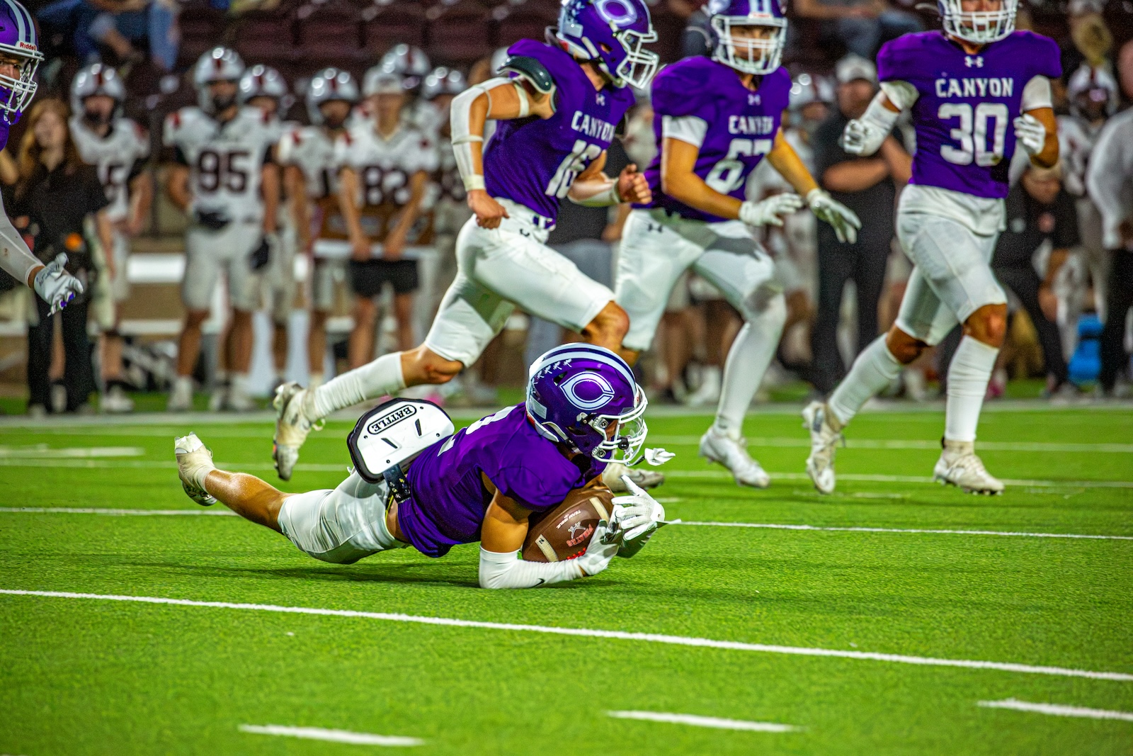 _V4A3423 | Press Pass Sports Purple-uniform football player lunges and secures a loose ball on bright green turf as teammates approach in the background.