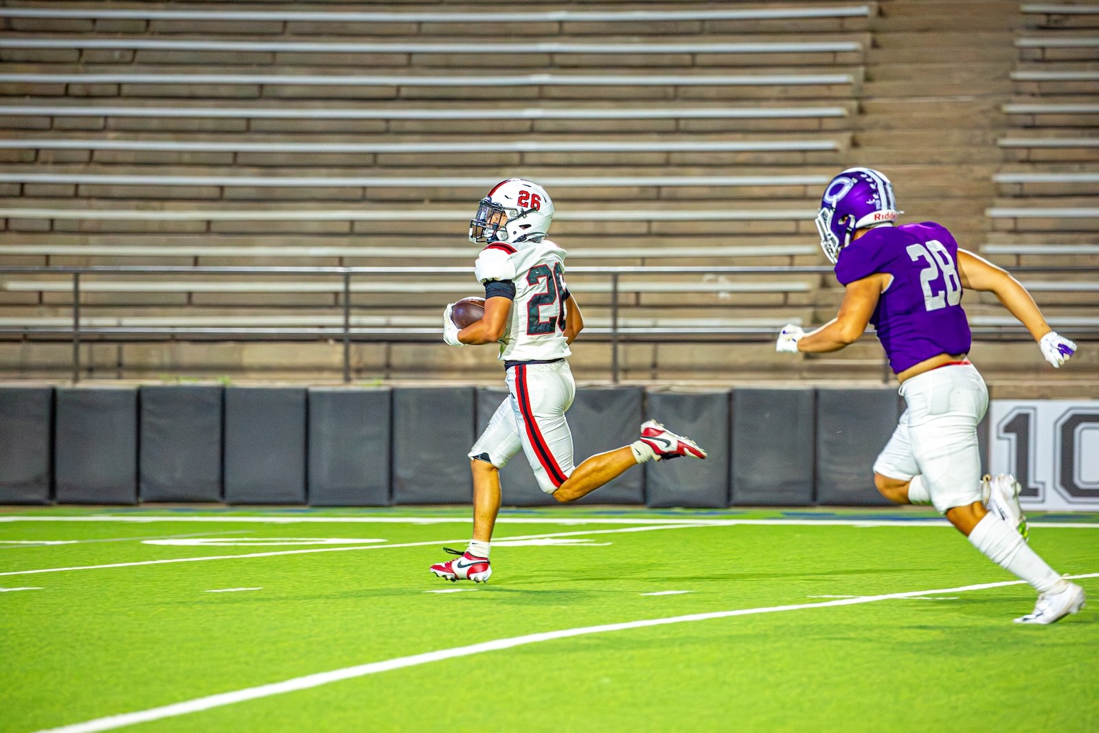 _V4A3537 | Press Pass Sports A football player in white with bib 21 runs with the ball while a defender in purple 28 pursues on a bright green turf field with empty bleachers in the background.