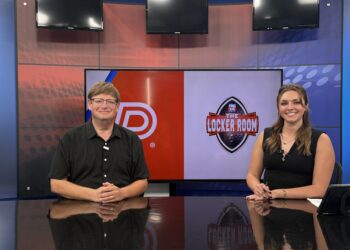 IMG_7477 | Press Pass Sports Two news anchors sit at a desk in a studio with a large screen behind them showing a red panel and a logo; man on left, woman on right smiling.