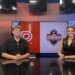 IMG_7477 | Press Pass Sports Two news anchors sit at a desk in a studio with a large screen behind them showing a red panel and a logo; man on left, woman on right smiling.
