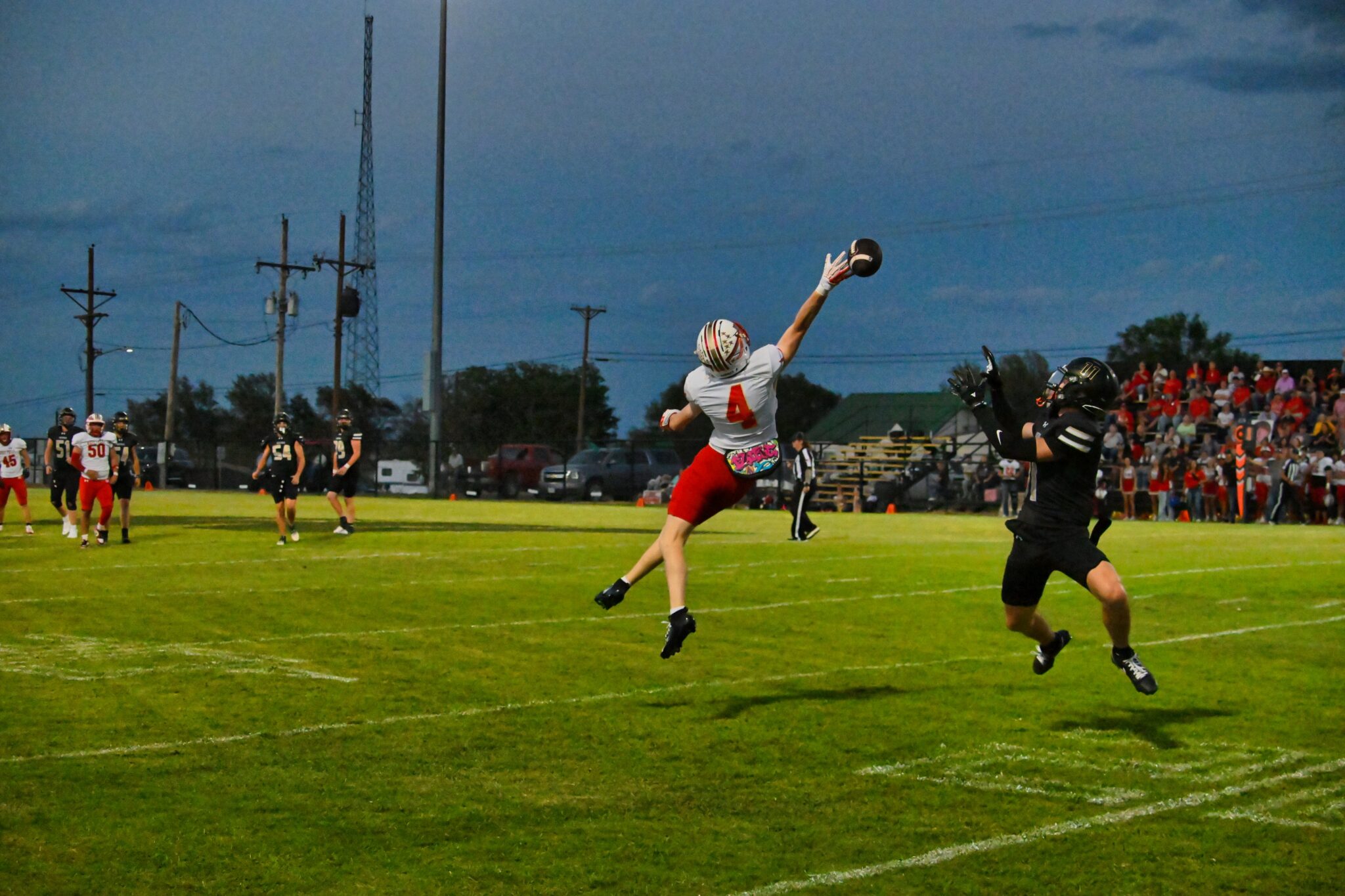 image0 34 | Press Pass Sports Football game action: a white‑jerseyed player leaps to catch a ball as a black‑jersey defender reaches in from the right on a grassy field at dusk, with a cheering crowd in the stands behind.