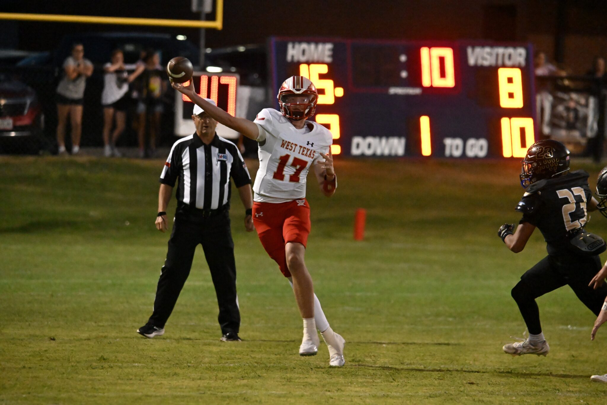 image1 26 | Press Pass Sports Football player in white West Texas jersey throws a pass as a referee watches on a night game, with a bright scoreboard in the background.