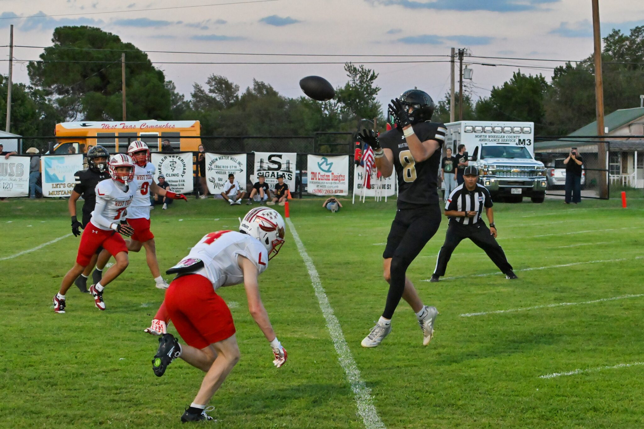image3 13 | Press Pass Sports Football players on a grassy field chase a spiraling ball as a receiver leaps to catch it, with banners and a parked ambulance in the background.