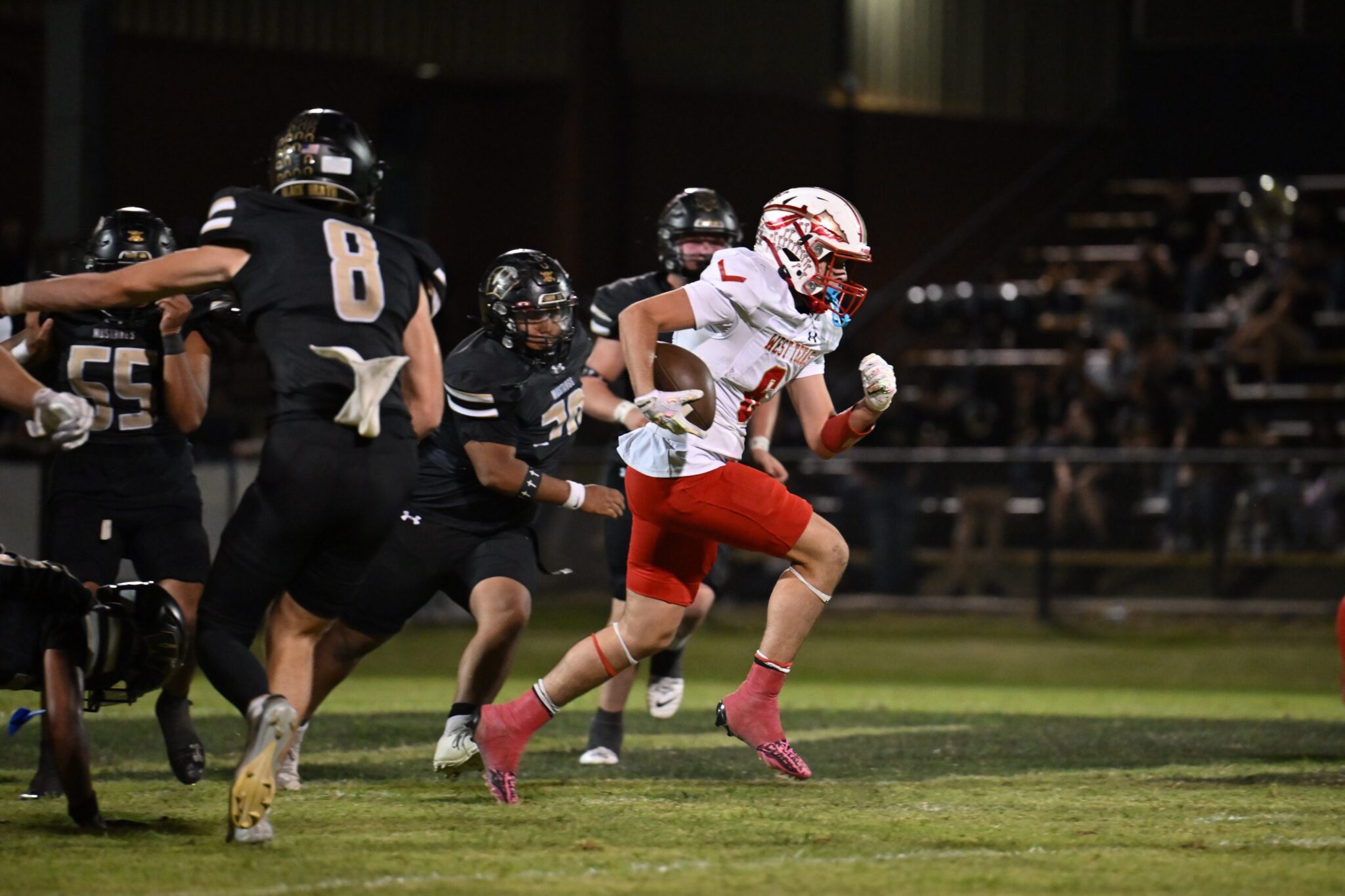 image4 3 | Press Pass Sports American football player in white and red carries the ball while pursued by several black-jersey defenders during a nighttime game on a grassy field, stadium in background.
