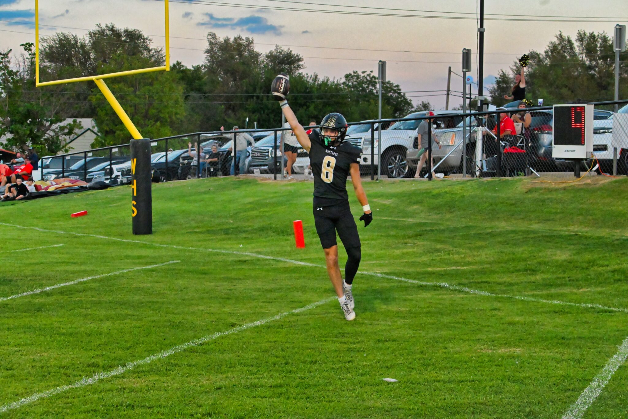 image4 4 | Press Pass Sports Football player in a black jersey with number 8 raises the ball in celebration near the end zone, with a yellow goalpost and cheering spectators in the background.