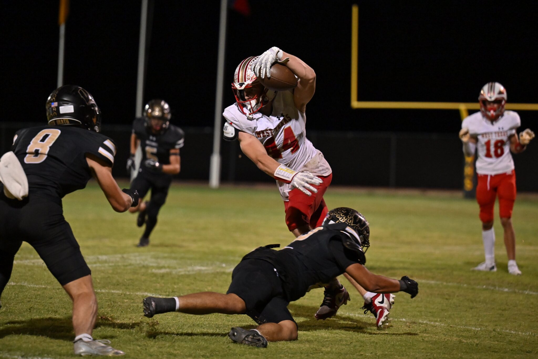 image5 5 | Press Pass Sports Nighttime football scene: a white-uniformed player cradles the ball while a defender on the ground reaches for him.