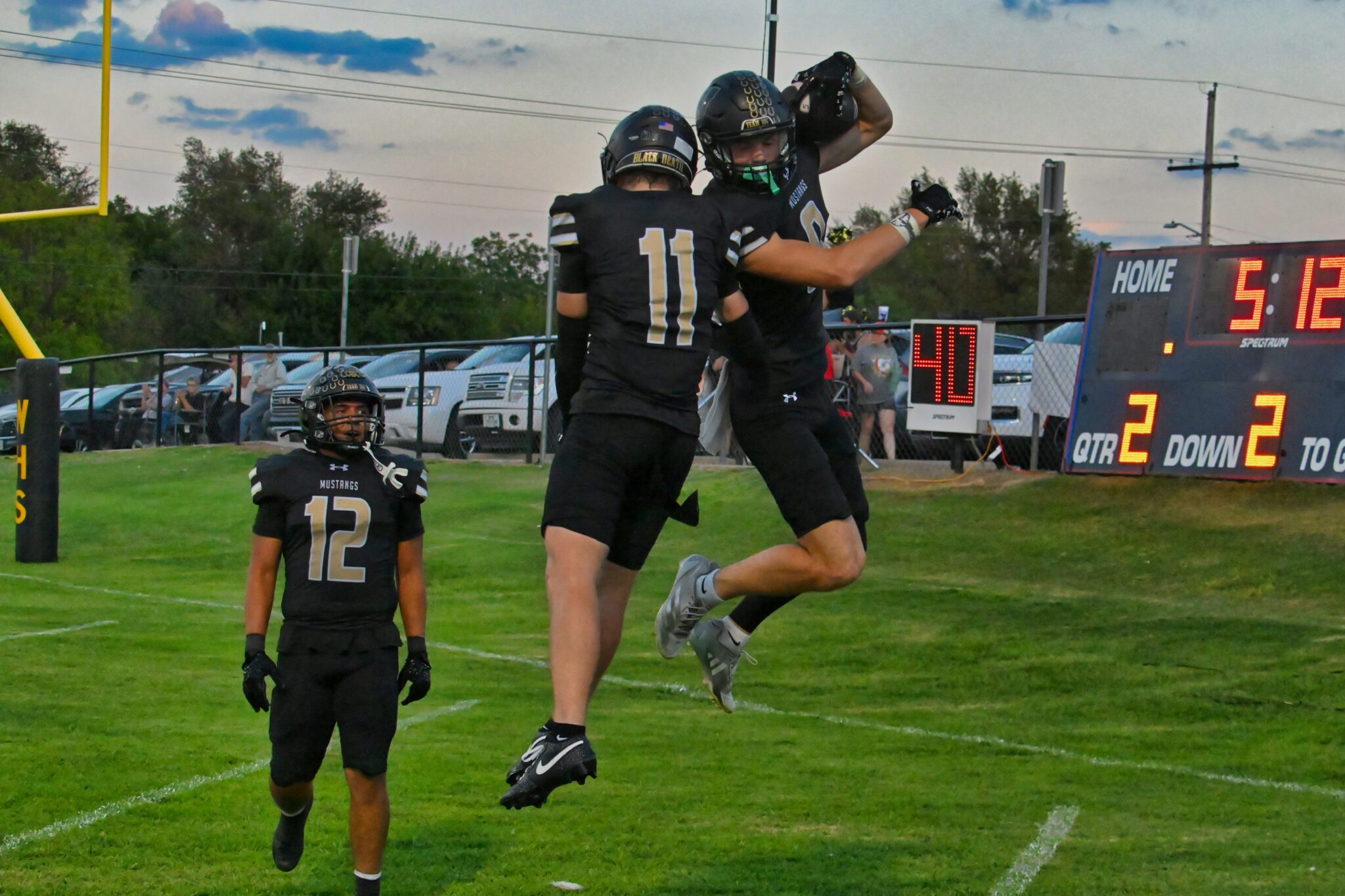 image5 6 | Press Pass Sports Two football players in black uniforms jump to celebrate near the end zone as a teammate watches, with a yellow goalpost and scoreboard in the background.
